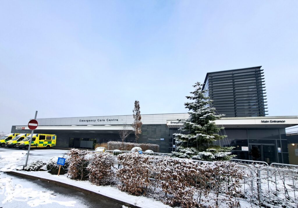 Wide angle photo of the front of the QE hospital in winter, with snow and a line of ambulances