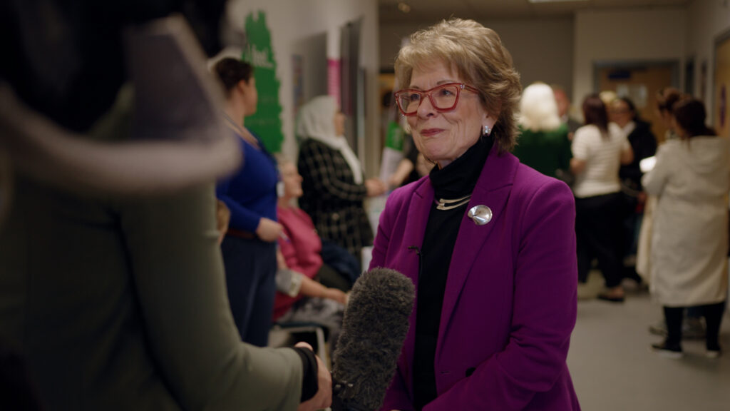 Photo of Dame Lesley Regan being interviewed with a back drop[ of a busy clinic