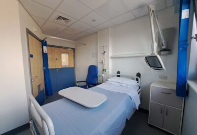 A photo of a single room on a hospital ward. There is a hospital bed, with a visitor's chair, a cabinet, TV screen and an over bed table.