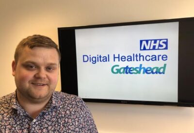 Head and shoulders shot of Adam Charlton with Digital Health Gateshead on a tv in the background