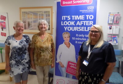 Brenda and Ann with the Breast Cancer Team stood next to a breast screening poster